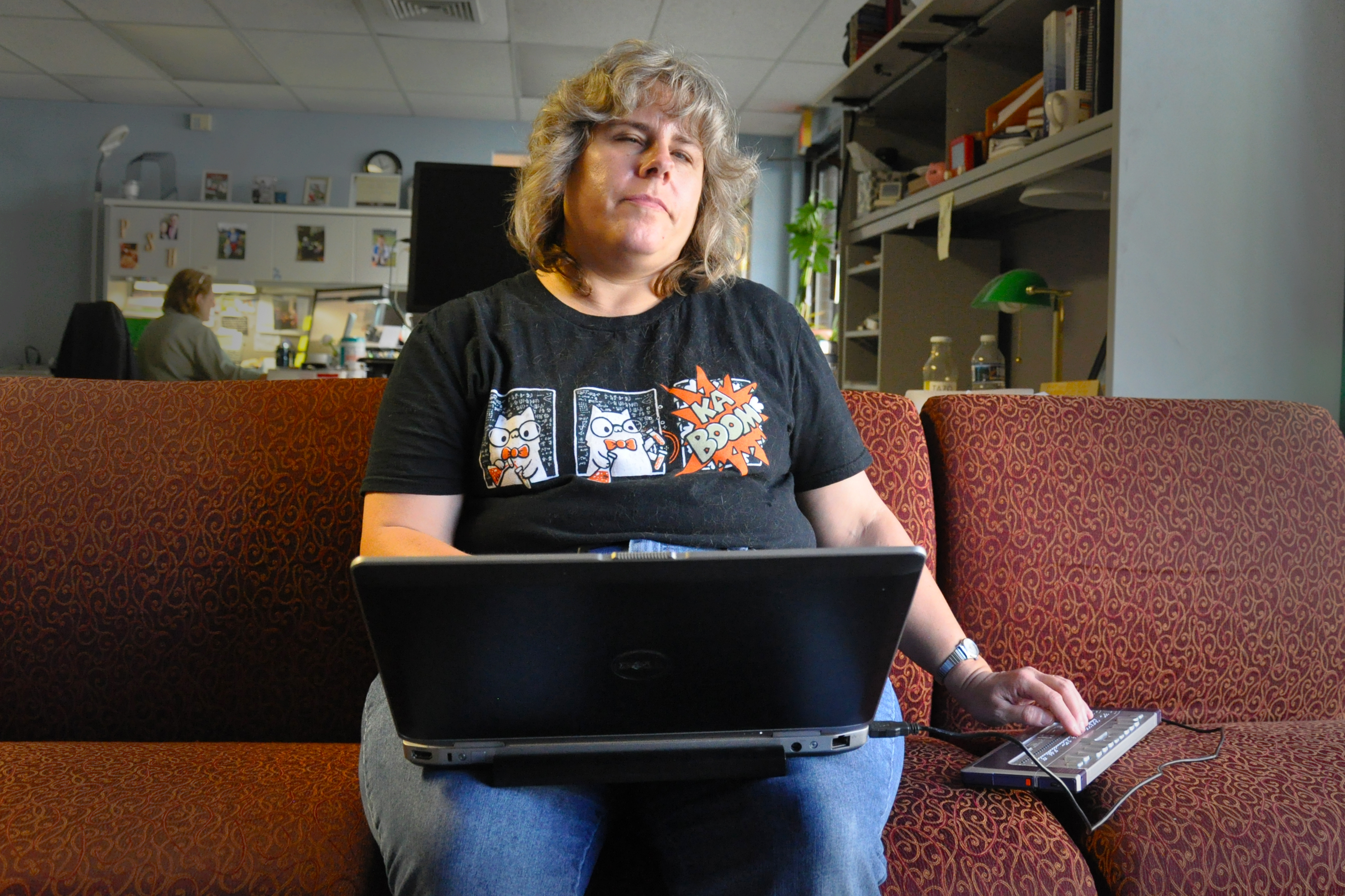 A woman sits on a couch with a laptop on her lap and a braille keyboard to her right