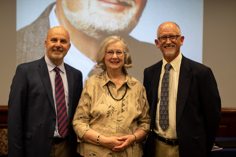 A woman stands between two men, all three of whom are smiling and facing the camera.