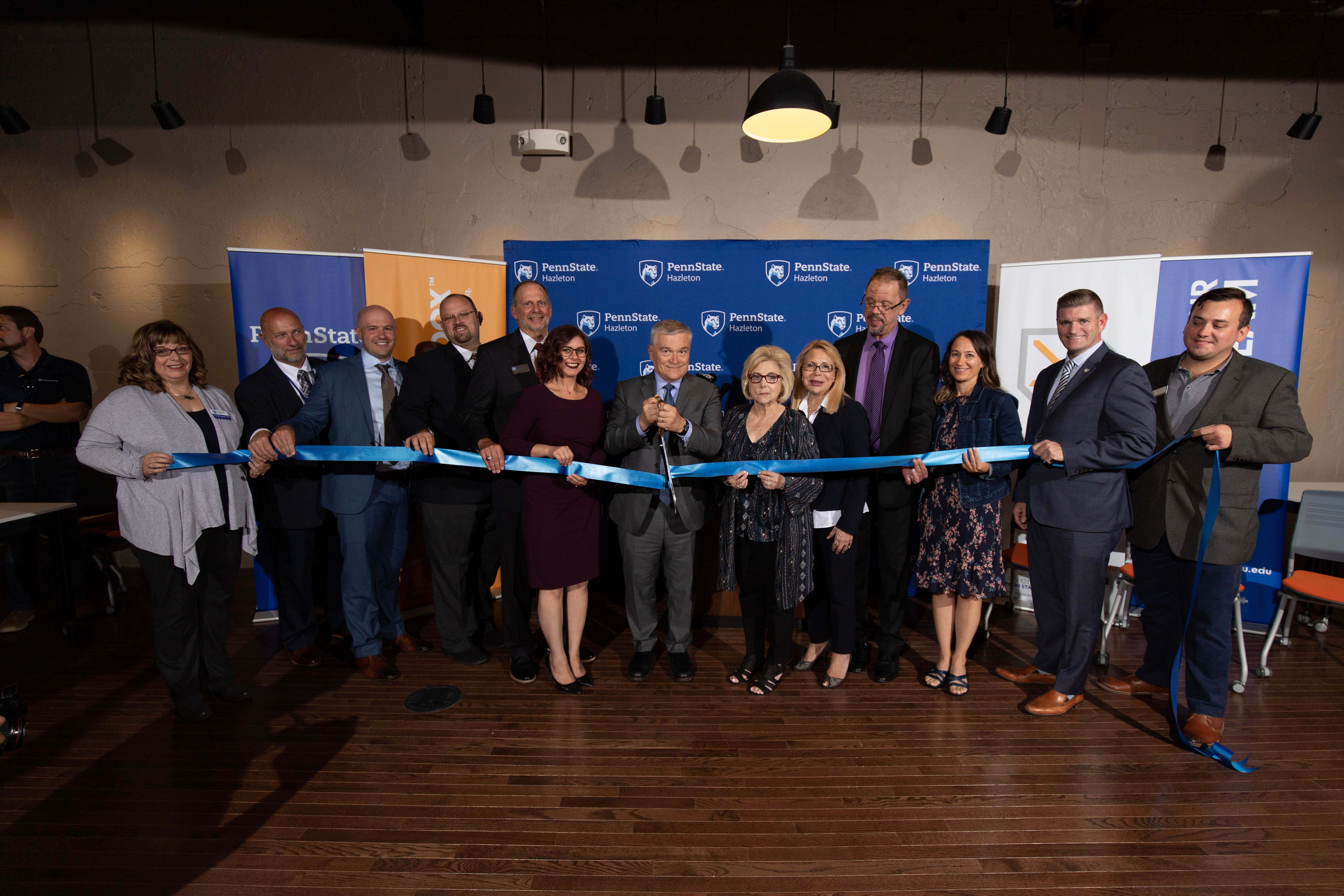 A group of eleven people lined up and holding a ribbon with one person cutting it. 