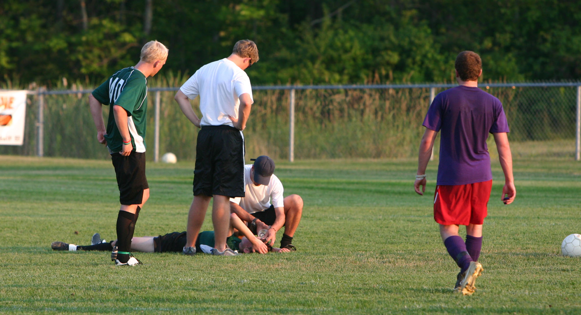 Injured soccer player laying on his back on the pitch. Concerned coaches attend to him as two players look on, concerned