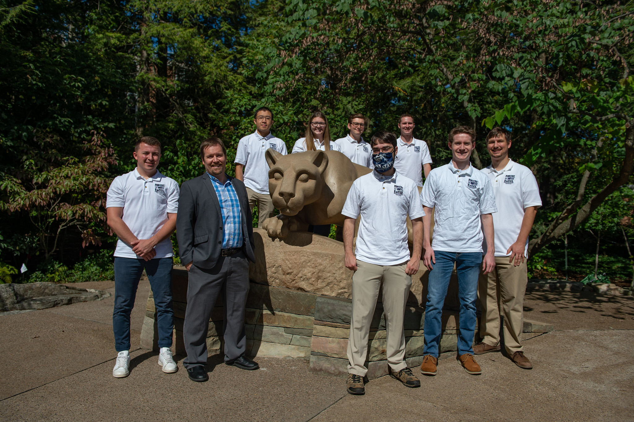 A professor and a group of students wearing white polo shirts pose in front of the Nittany Lion shrine. 