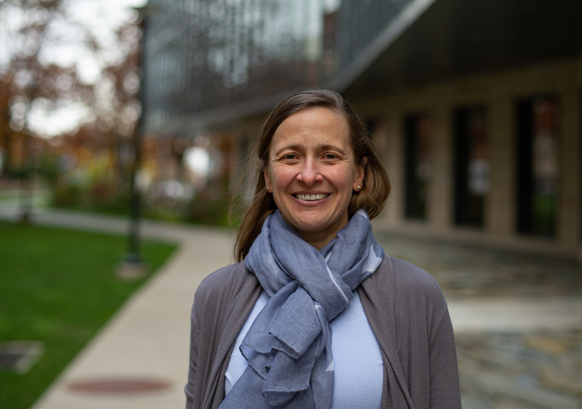 A researcher wearing a gray cardigan and blue scarf smiles with an engineering building and sidewalk in the background