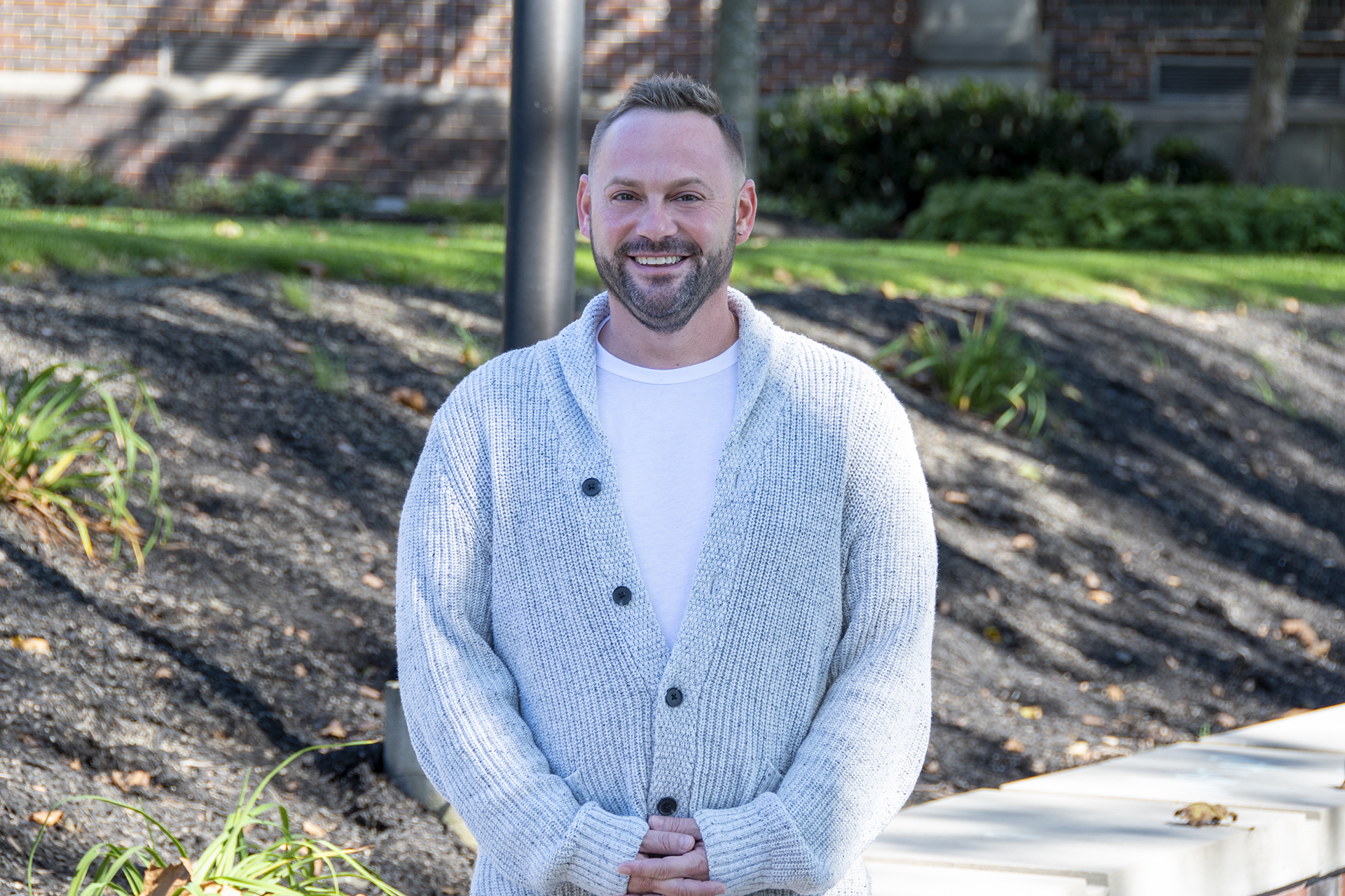 Smiling man wearing a white shirt and cardigan stands with hands clasped in front of him.