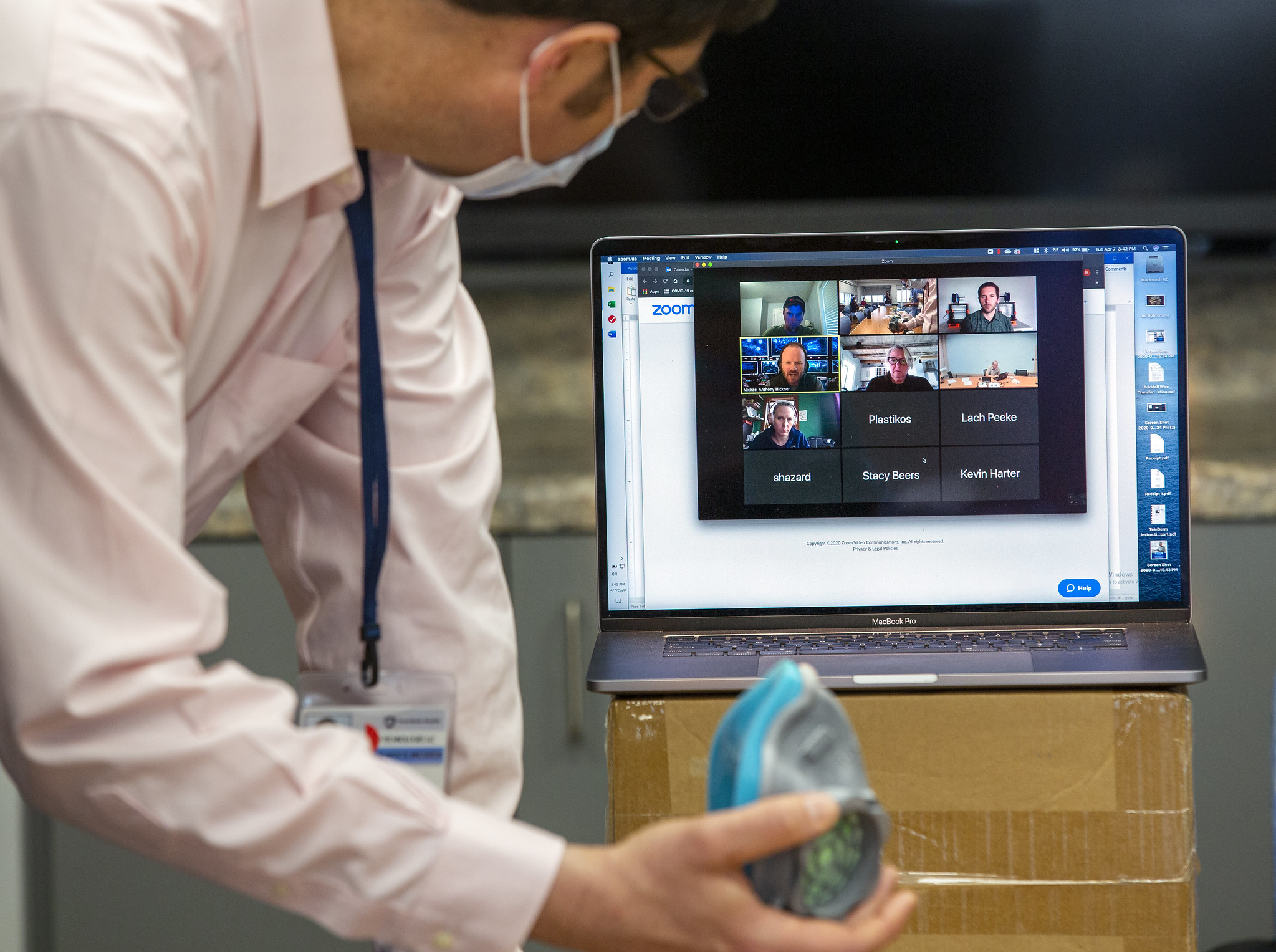 A man holds a 3D printed N95 respirator while looking at a computer screen on which several people are visible as part of a Zoom meeting.