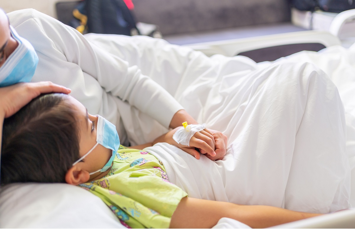 Young mother with protective face mask holding son's hand while lying on the hospital bed with him.