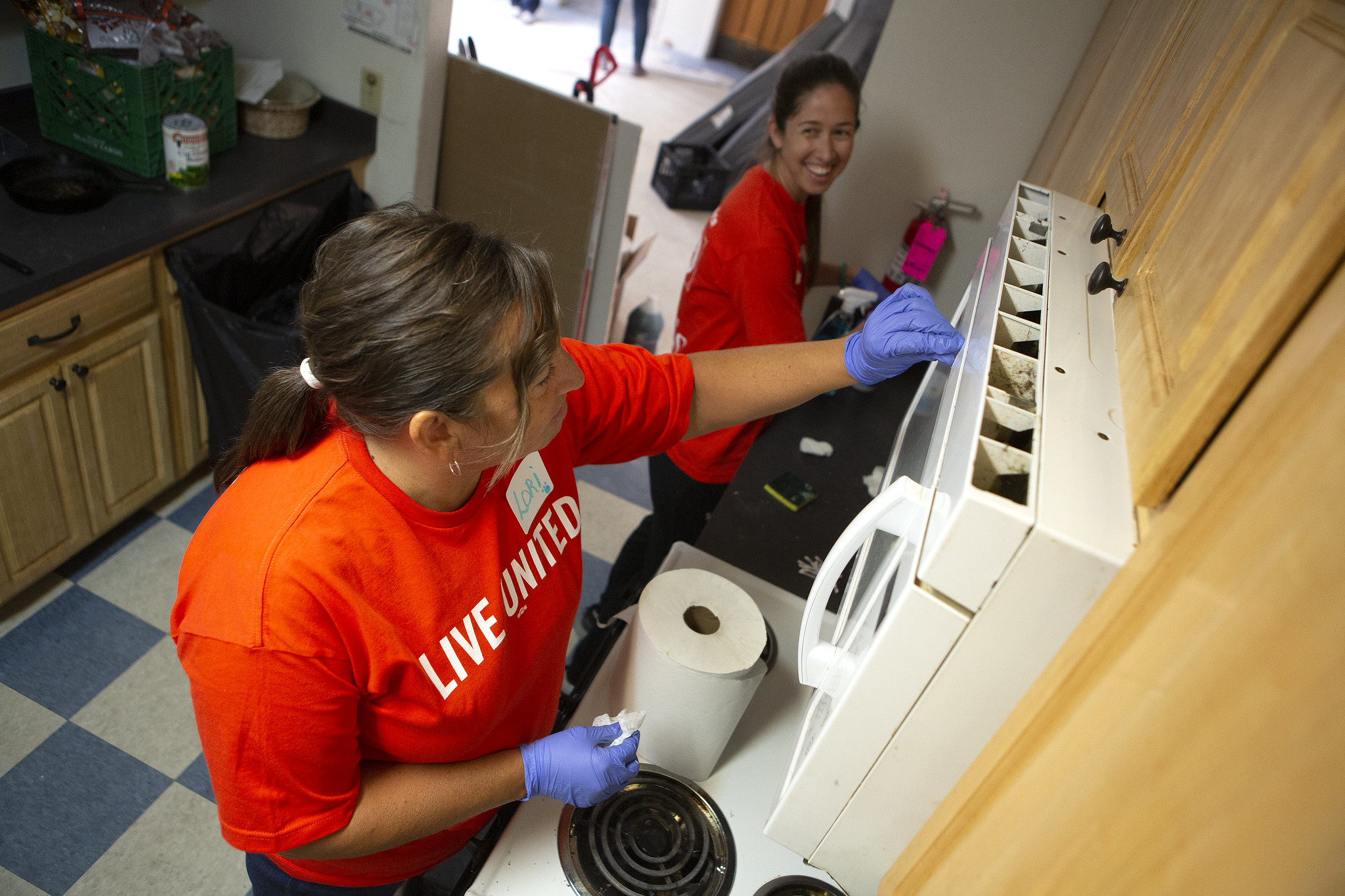 Two women wearing rubber gloves work side-by-side to clean a kitchen in a residential apartment. One looks to the other, smiling.