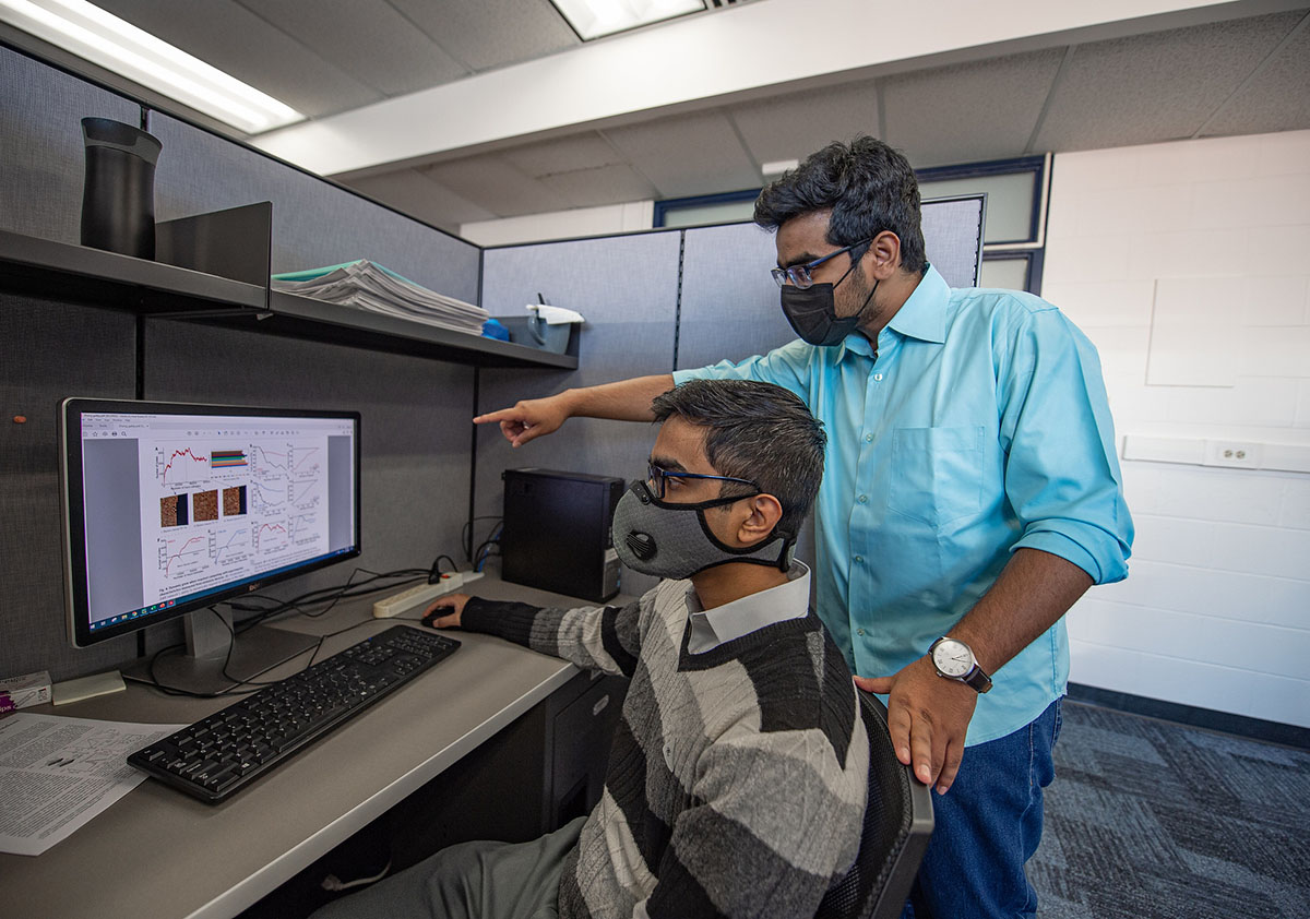 A person sits in front of a computer displaying graphs while another person stands behind him and points. 