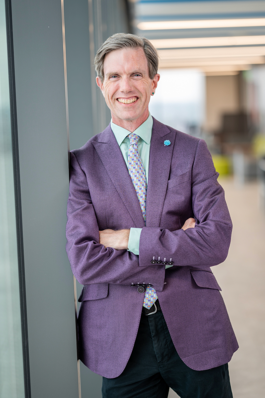 white male in purple suit leaning against window in hallway