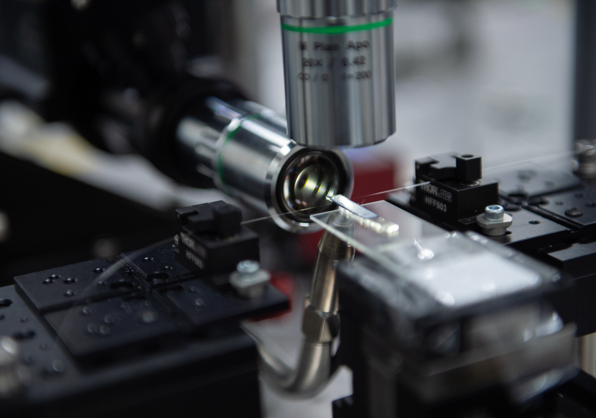 A glass rectangle sits at the forefront, slightly out of focus, touching a hair-like structure stretched between two black clasps. Two silver cylinders point at the structure, one from above and one across from the glass rectangle. 