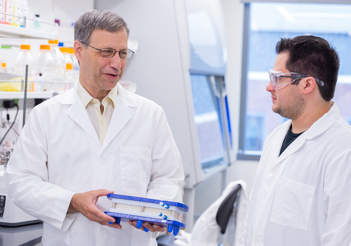 A man smiles and holds a membrane model while a male student in a lab coat looks on.