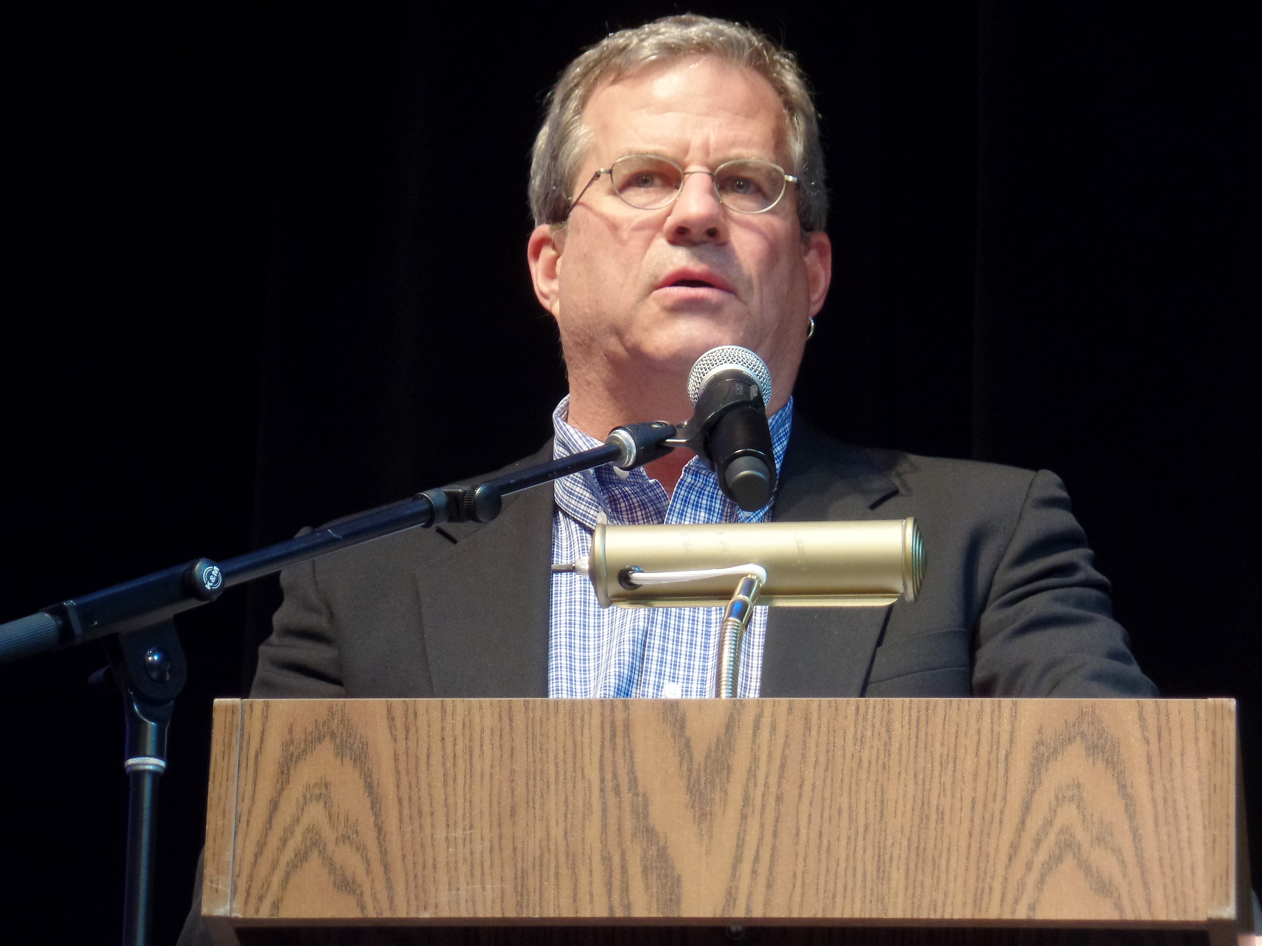 Sam Quinones, a white man with grey hair speaking into a microphone at a podium