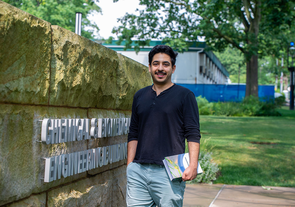 A person wearing a dark long sleeved shirt holds a textbook in front of a "Chemical and Biomedical Engineering Building" sign on a stone wall outside