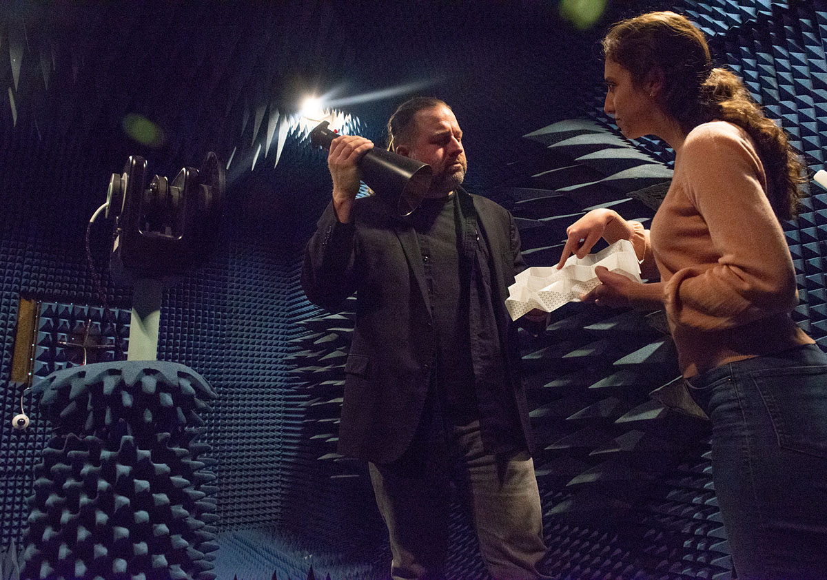 Two researchers stand in an anechoic chamber. One holds a conical horn antenna and one holds an origami frequency selective surface. 