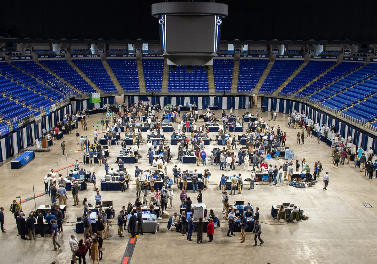 Photo taken from stands shows nearly eighty display tables set up in a large arena surrounded by people presenting and talking.