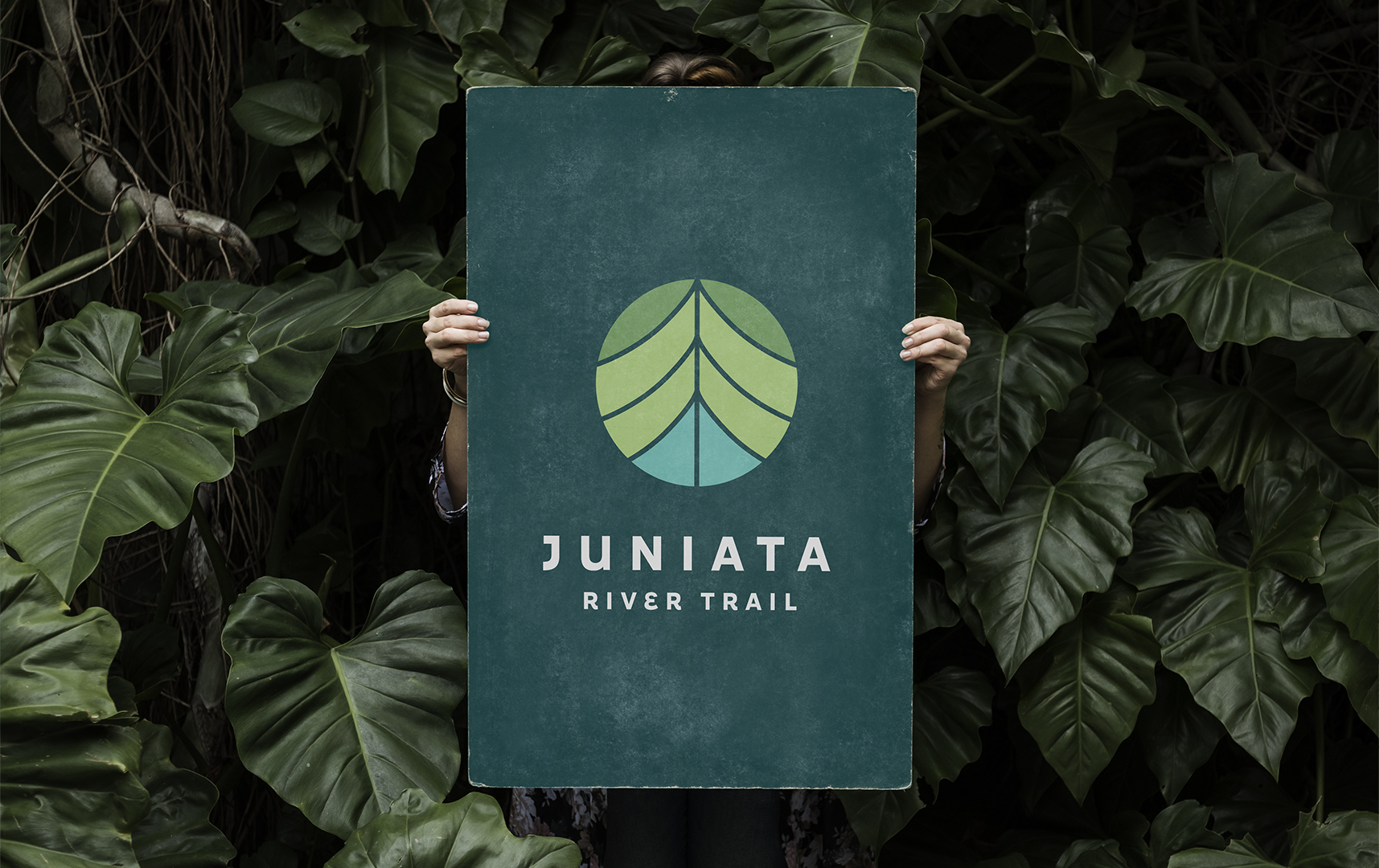 Someone holding up a sign for the Juniata River Trail in front of their body among a background of plants.