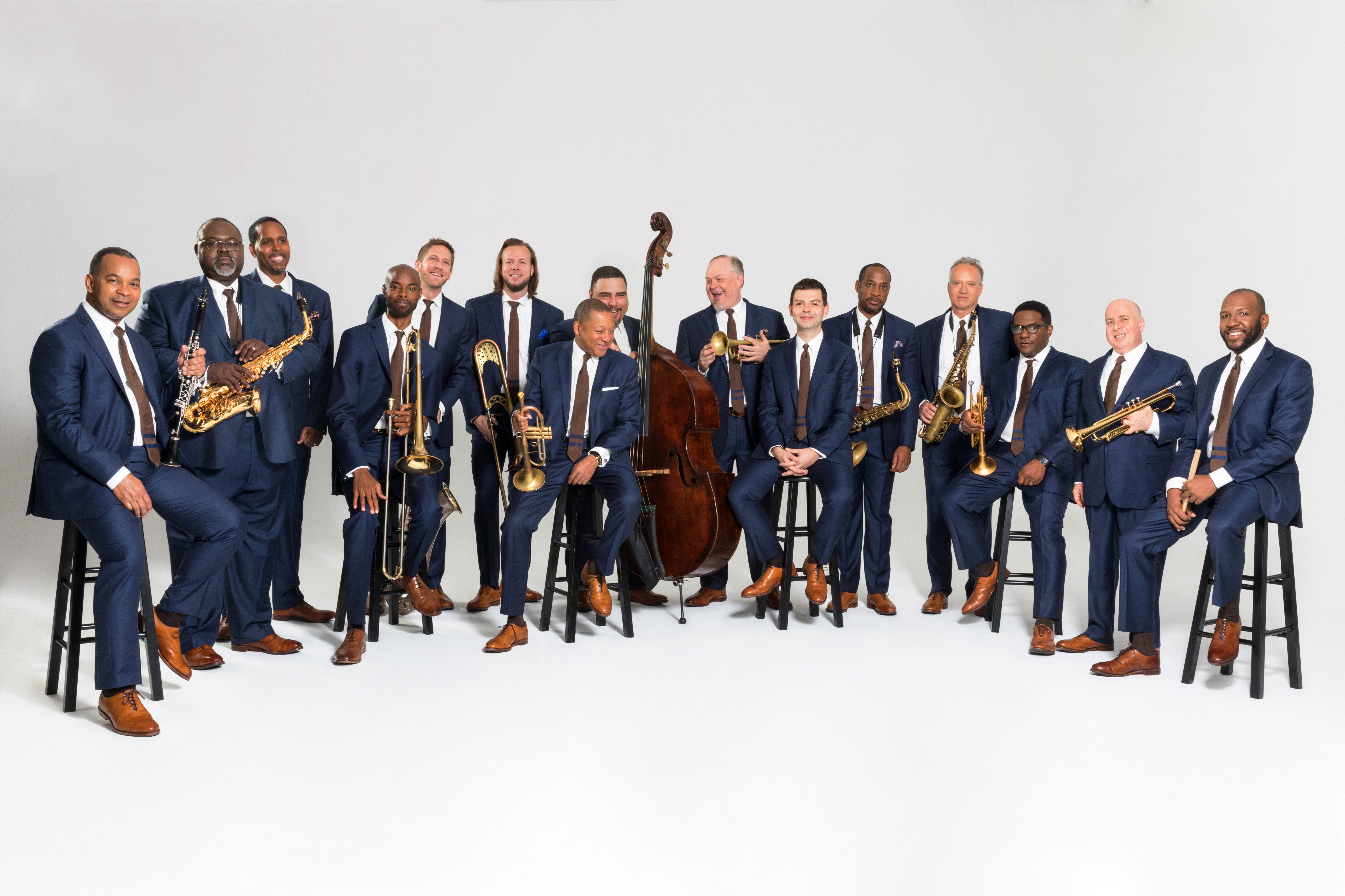 A large group of men all wearing suits stand and sit on stools while they hold their instruments.