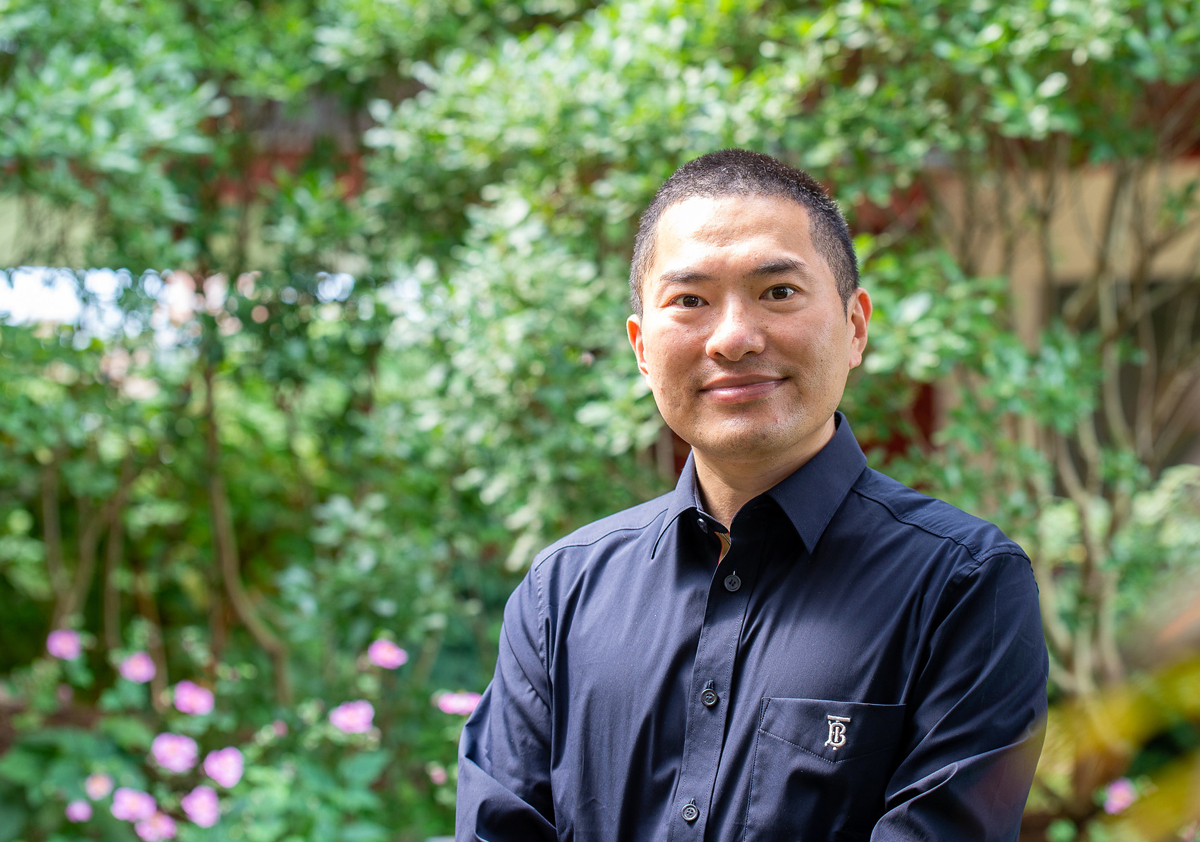 A researcher wearing a black button-down shirt smiles for a portrait in front of greenery.