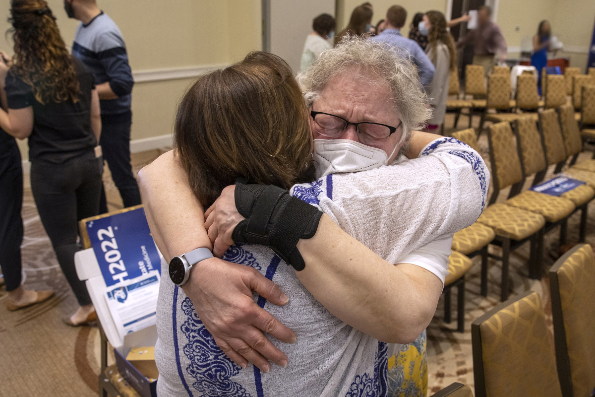 Two women embrace. The woman facing the camera has her eyes closed. Several people standing and chairs are in the background.