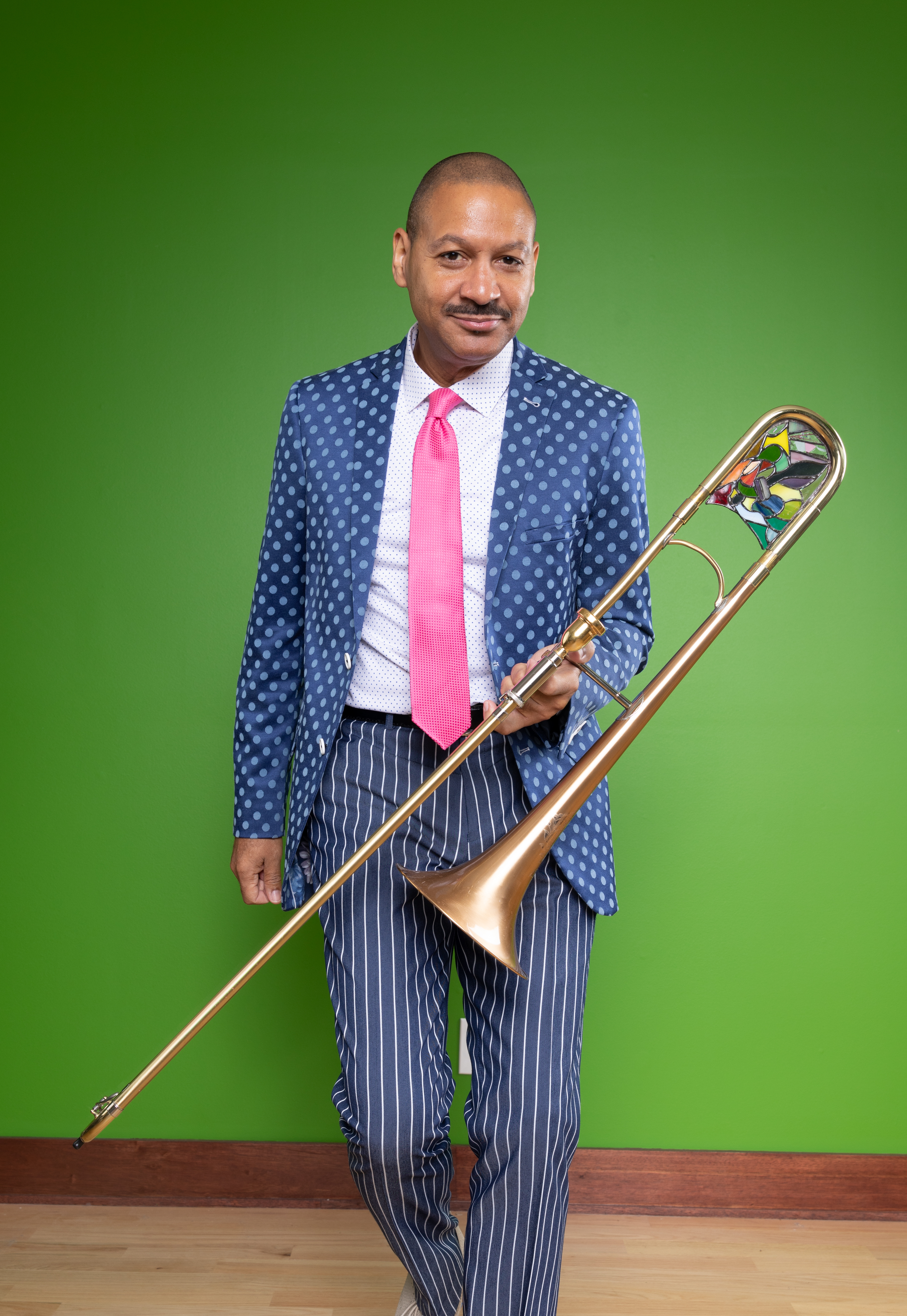 A man wears a suit and leans against a wall while he holds a trombone.