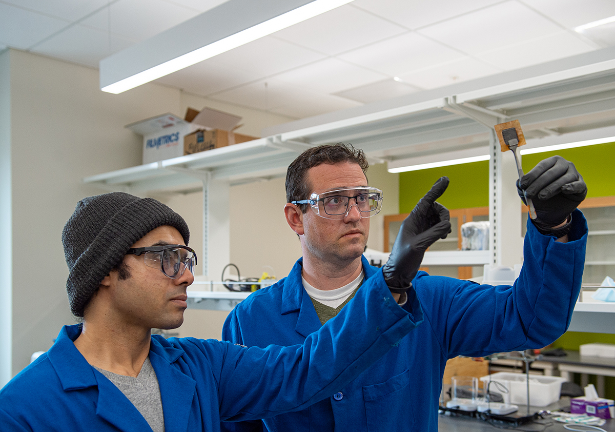 Two men in lab coats and protective glasses look up at a yellow, square membrane sample held by a lab tool resembling tweezers.