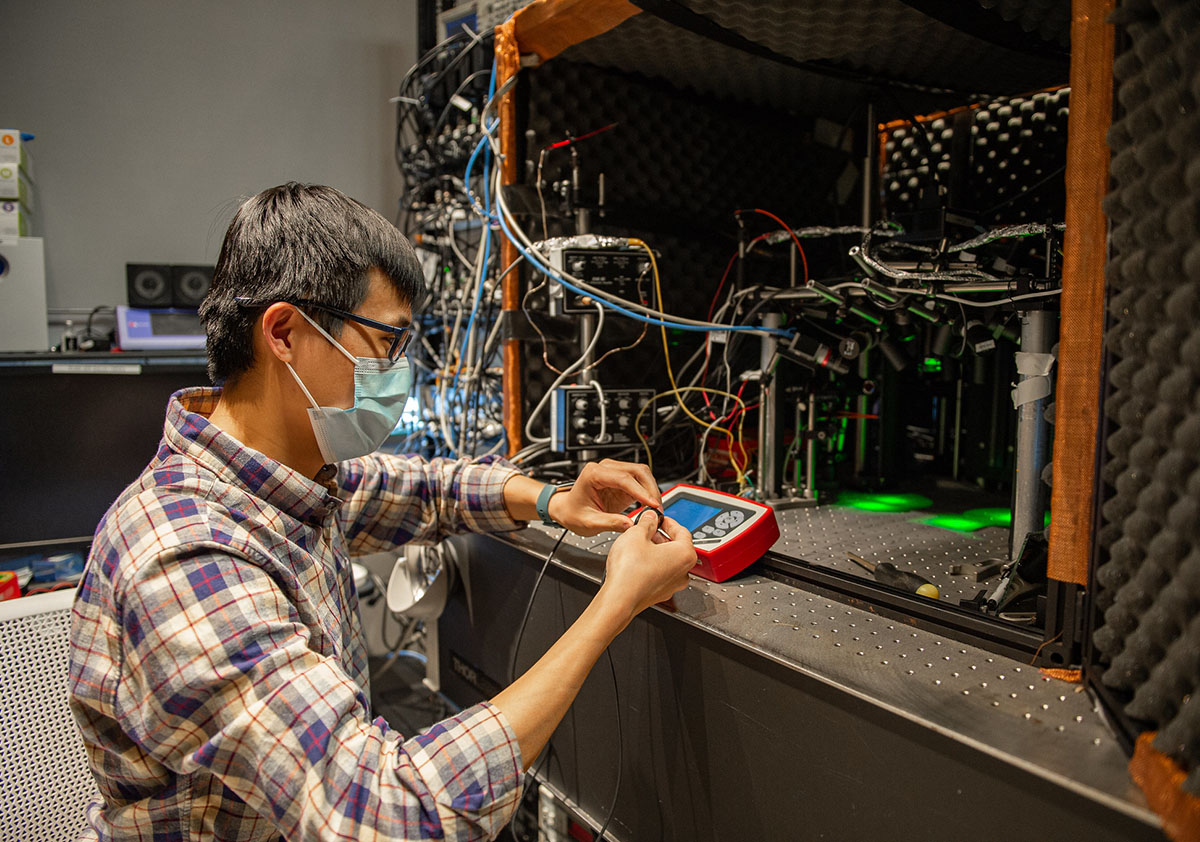 A researcher in a plaid shirt and blue face mask adjusts a set of wires in a lab setting.