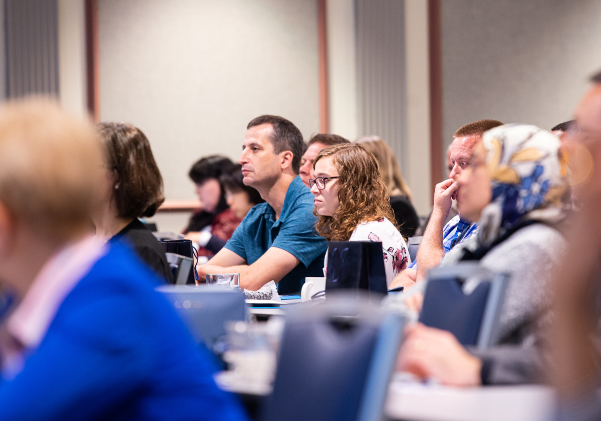 Many people sit in a lecture hall with laptops looking toward the front of the room