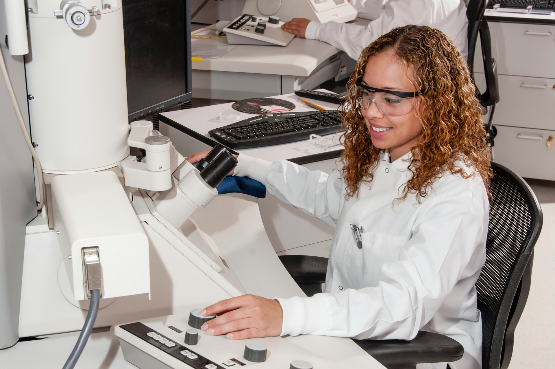 A person in a white lab coat and safety glasses works at a lab station.