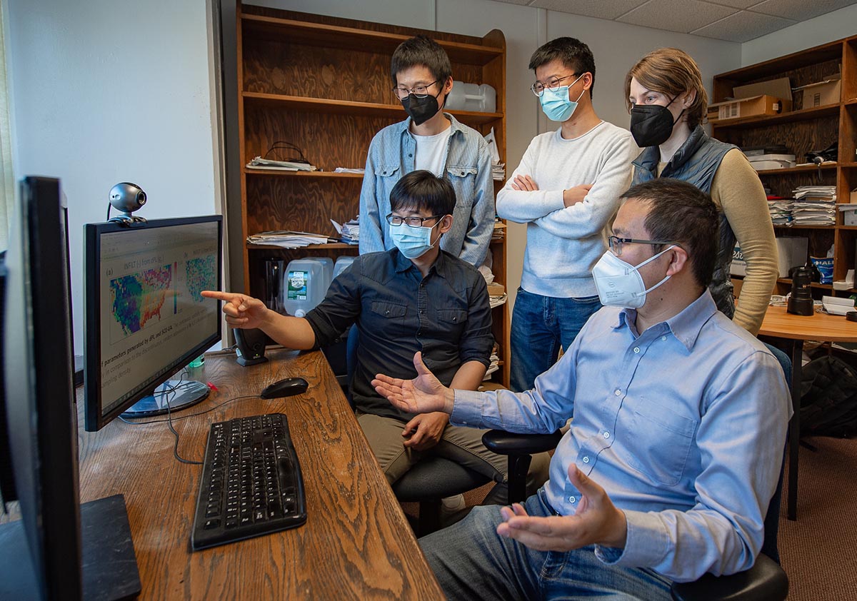 Five researchers wearing masks stand and sit around a computer, where two maps of the United States are displayed on the monitor. 