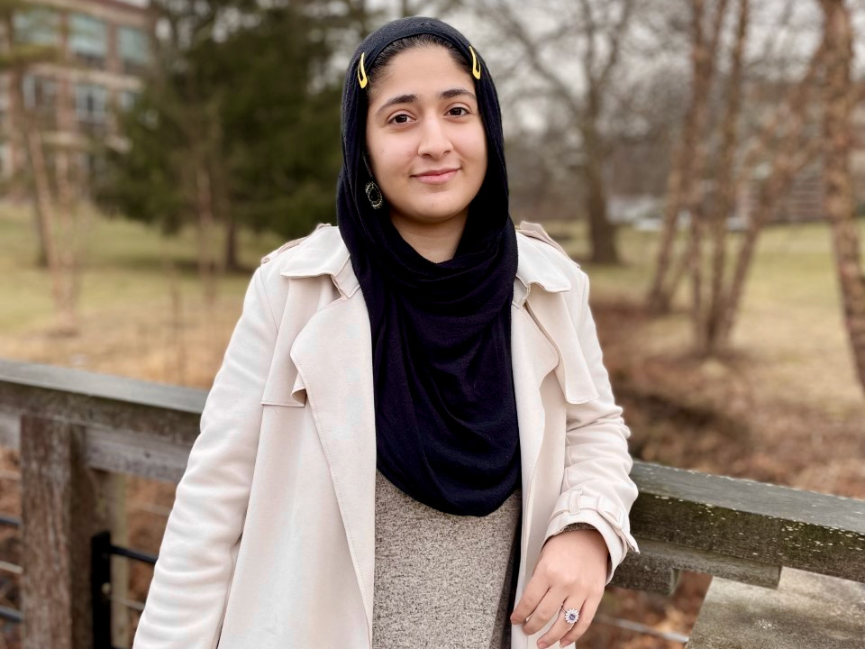 Rabia Rafiqzadah stands on a bridge wearing a black scarf and white coat. 