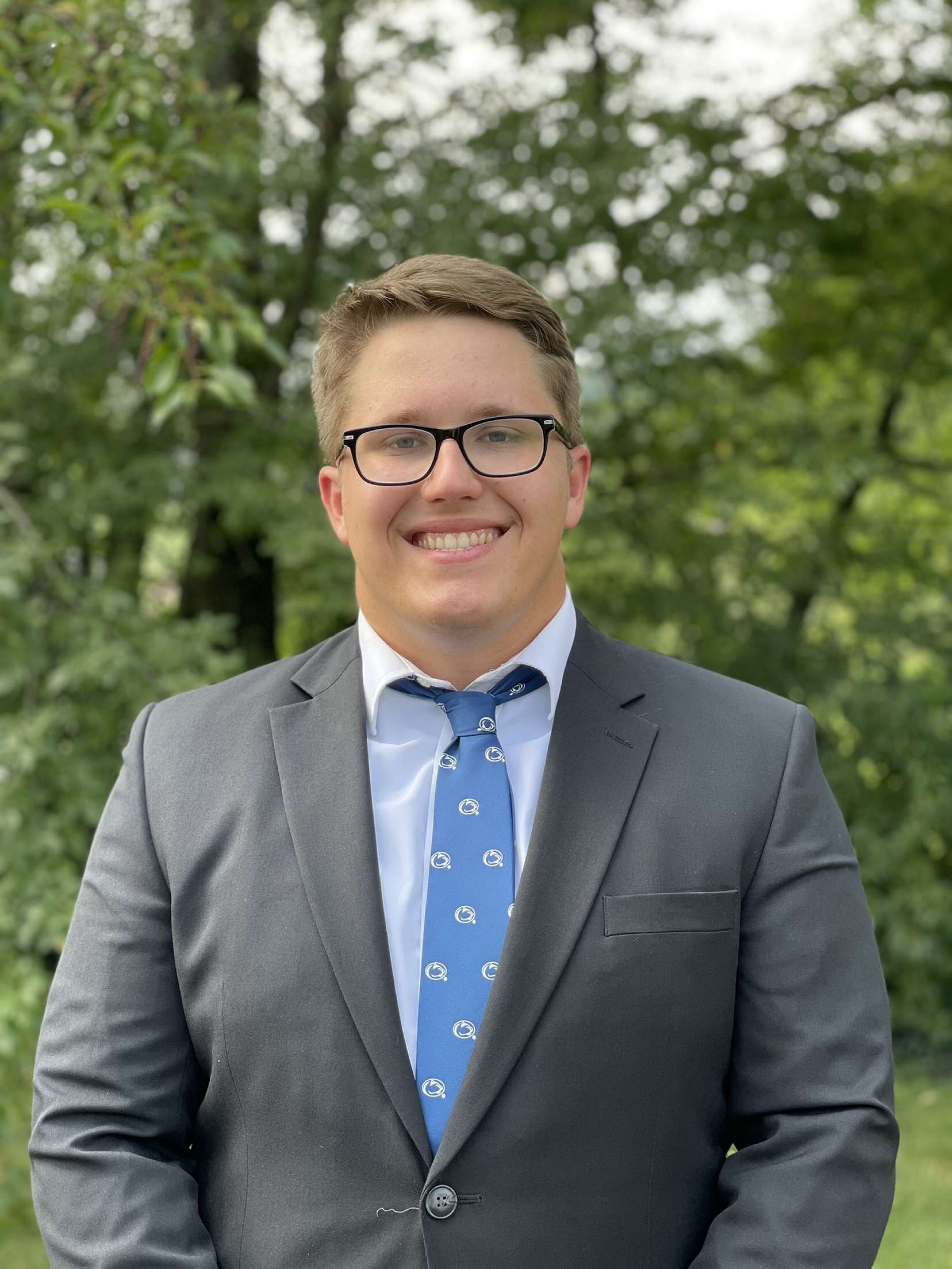 A male student smiling and wearing a suit and tie with trees in the background