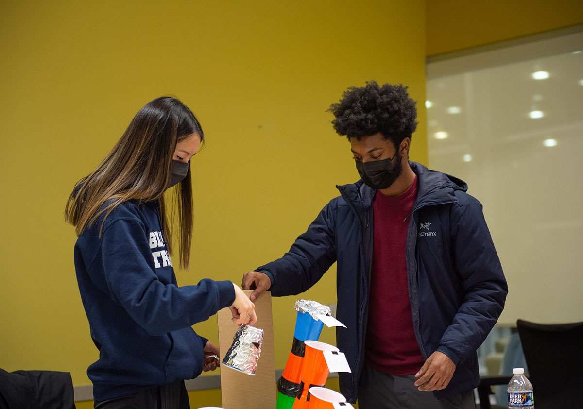 Two students stand in front of a yellow wall and arrange plastic cups and other materials.