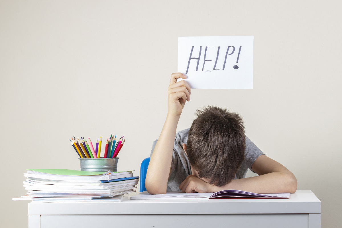 Sad tired frustrated boy sitting at the table with many books and holding help sign.