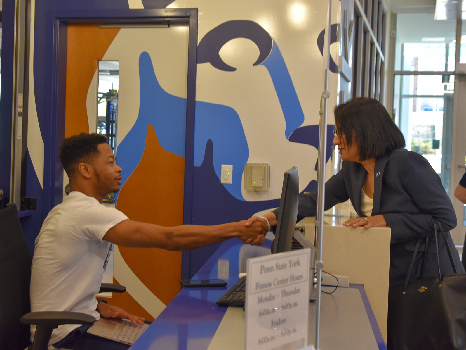 Male student of color seated at computer desk reaching to shake hand with a dark-haired woman eearing glasses.