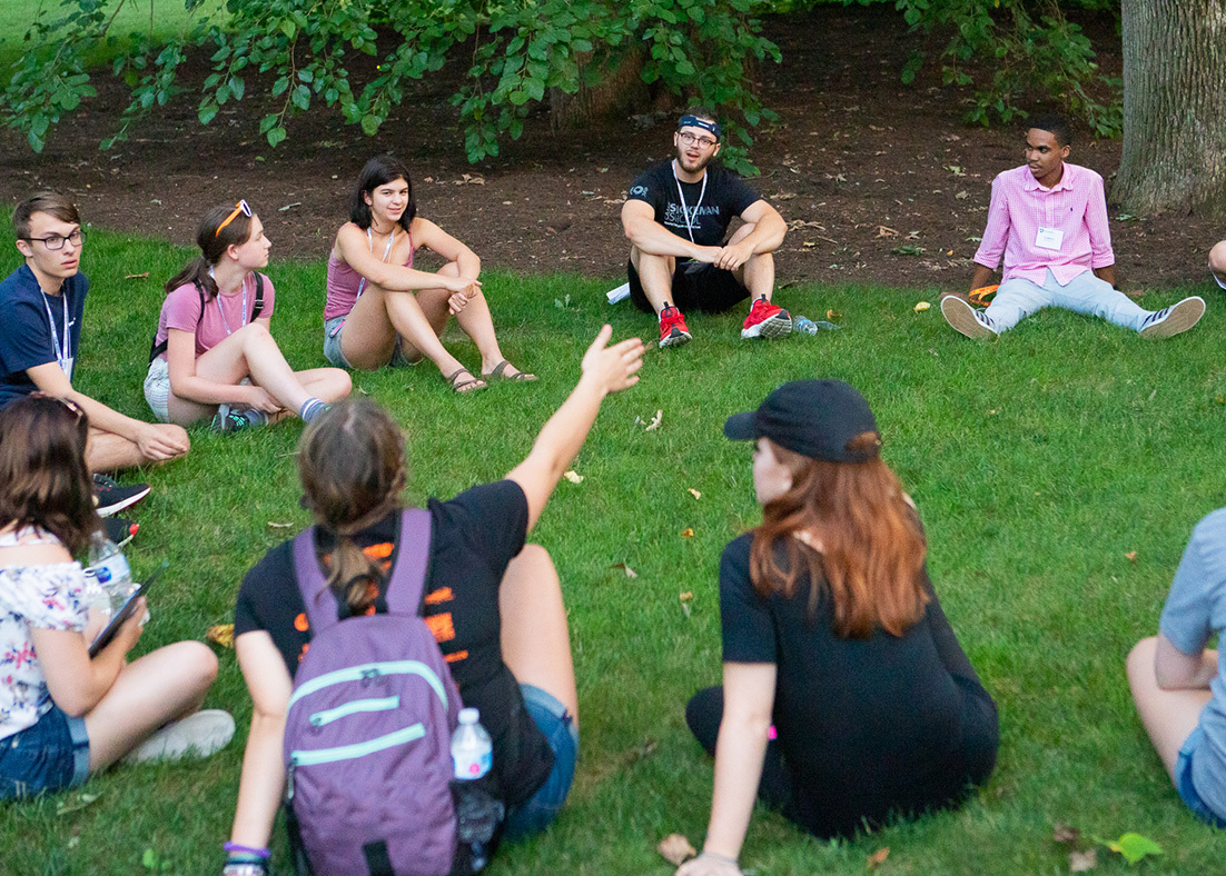 Summer camp students sitting in a circle on a patch of grass and talking.