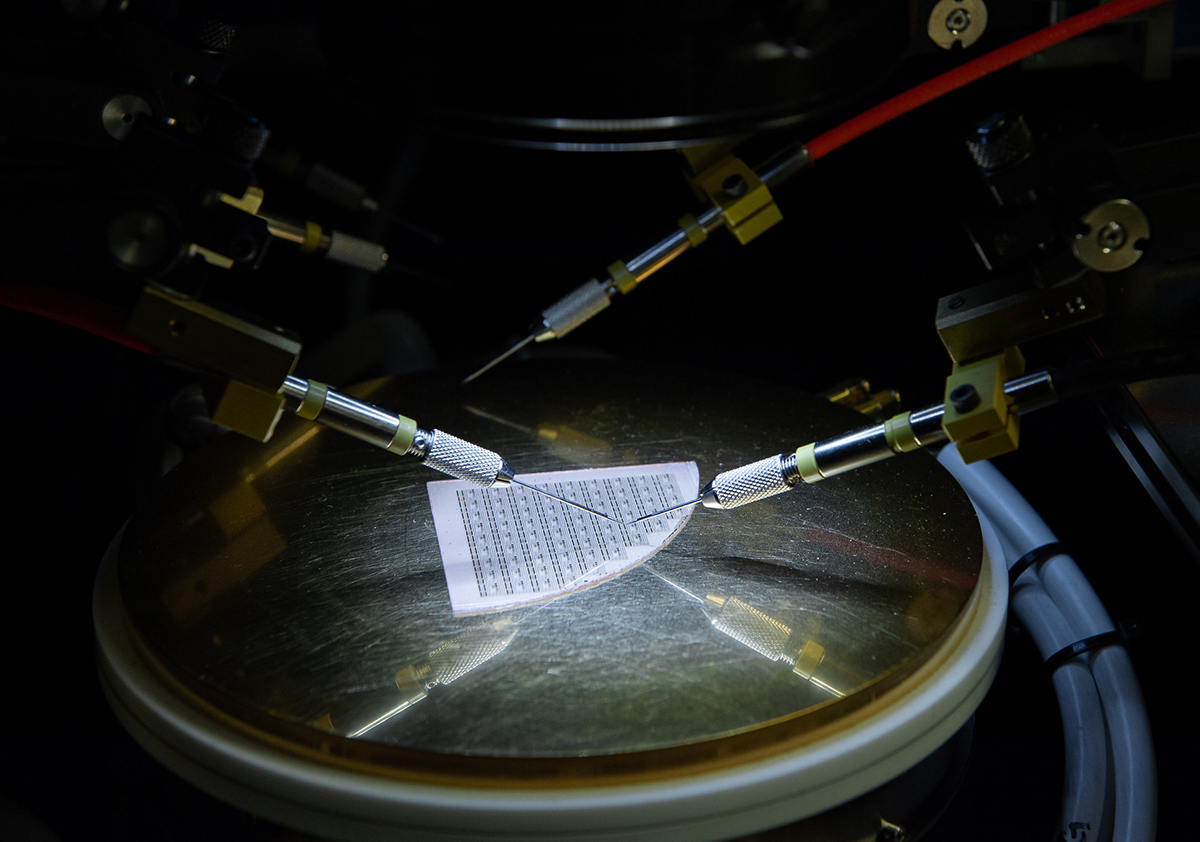 A white square with black lines sits on a metal circle, surrounded by three thin needles attached to larger machinery. 