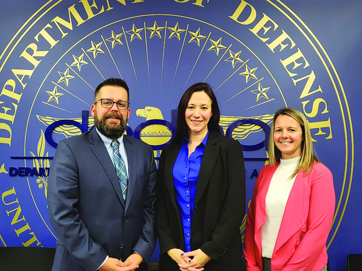 Three individuals stand against a blue wall with a gold Department of Defense seal