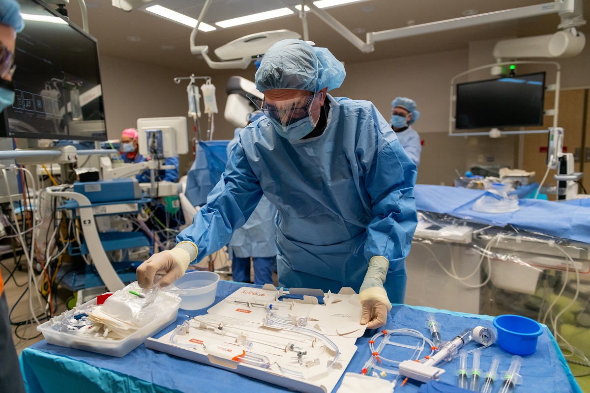 A man in hospital scrubs, a mask and gloves prepares surgical instruments on a table in an operating room. Behind him, other health care professionals work around instruments.