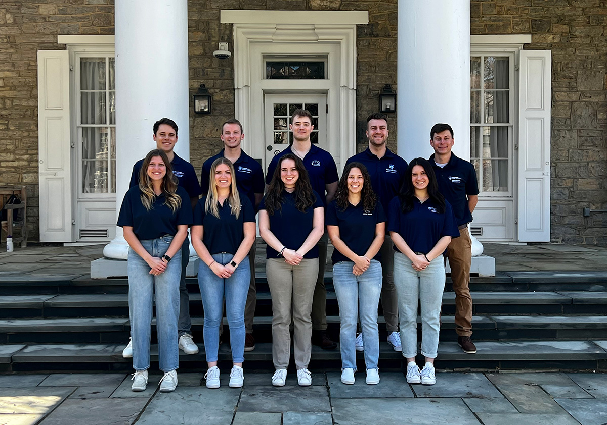 Two rows of five students wear matching blue shirts and stand on steps in front of a beige building. 