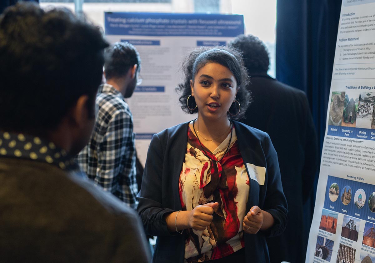 A woman stands in front of a large poster and explains her research to a person in the foreground.