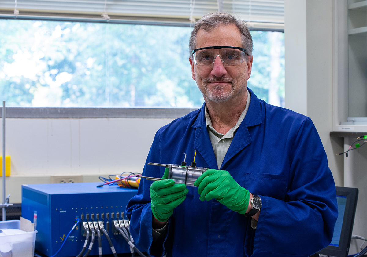 A man in a blue lab coat and protective glasses holds a small electrochemical reactor.