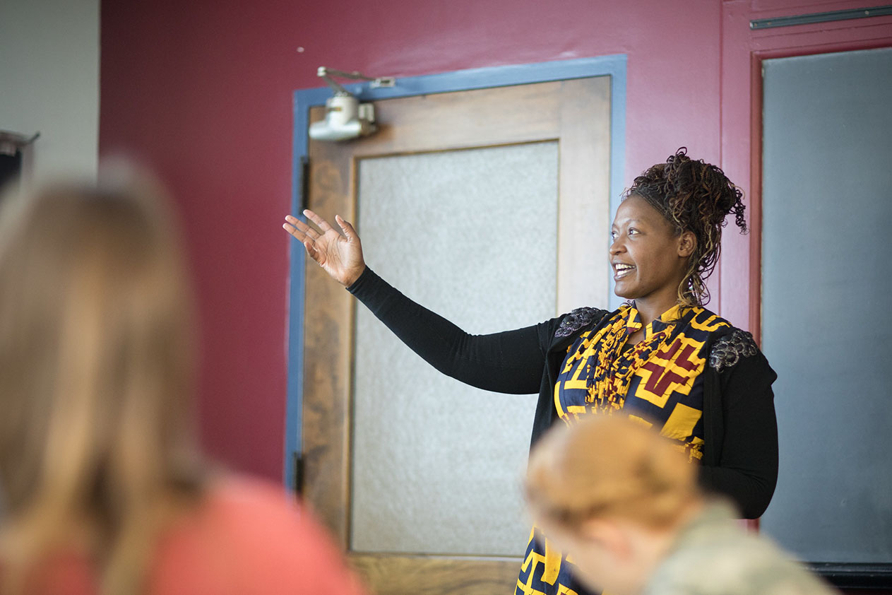 A woman wearing a black and yellow top gestures at a chalkboard while two students in the foreground look on. 