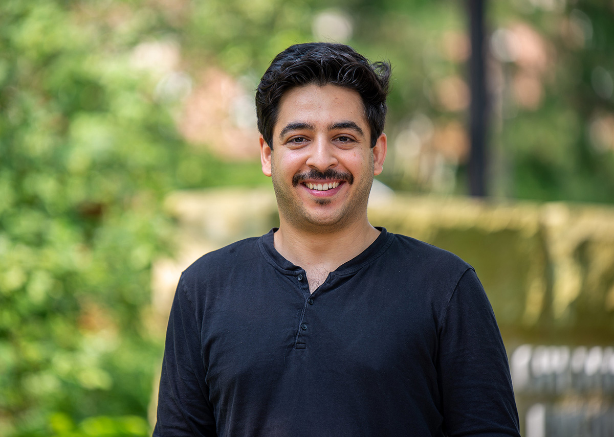 A man smiles at the camera. Greenery is in the background. 