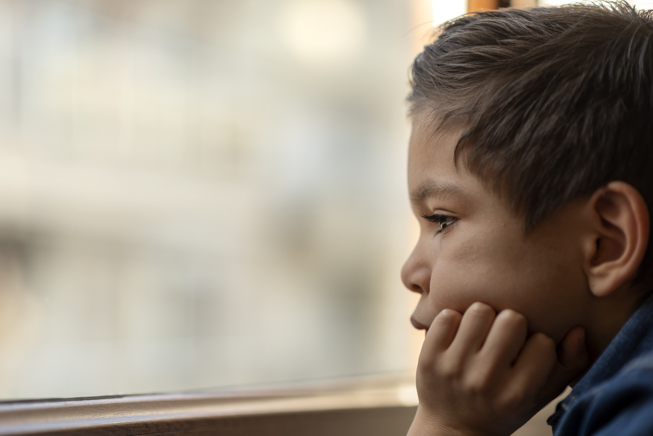 A young boy sits with his head in his hand, looking out a window