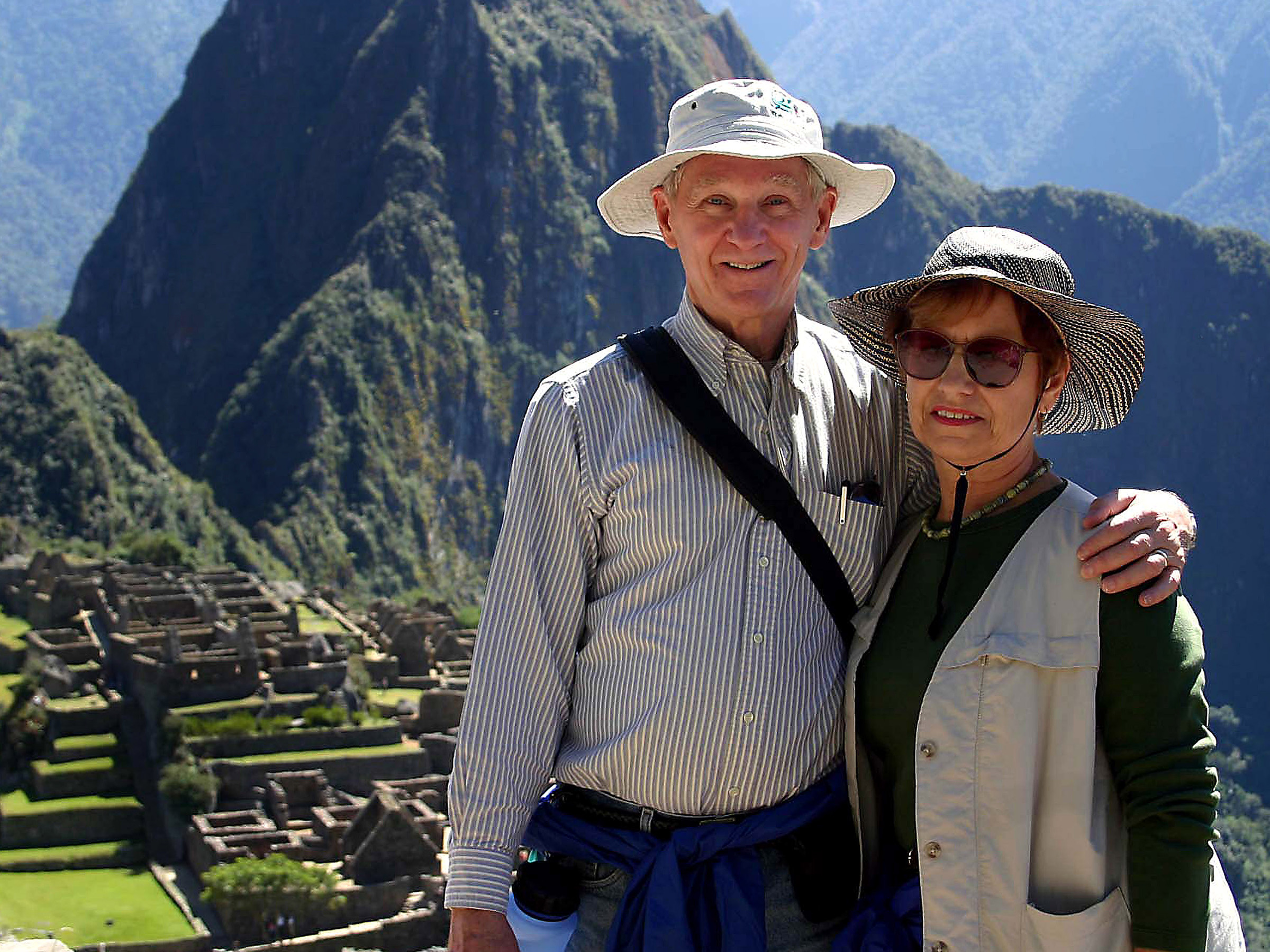 A man and woman wearing sun hats pose on Machu Picchu mountaintop in Peru.