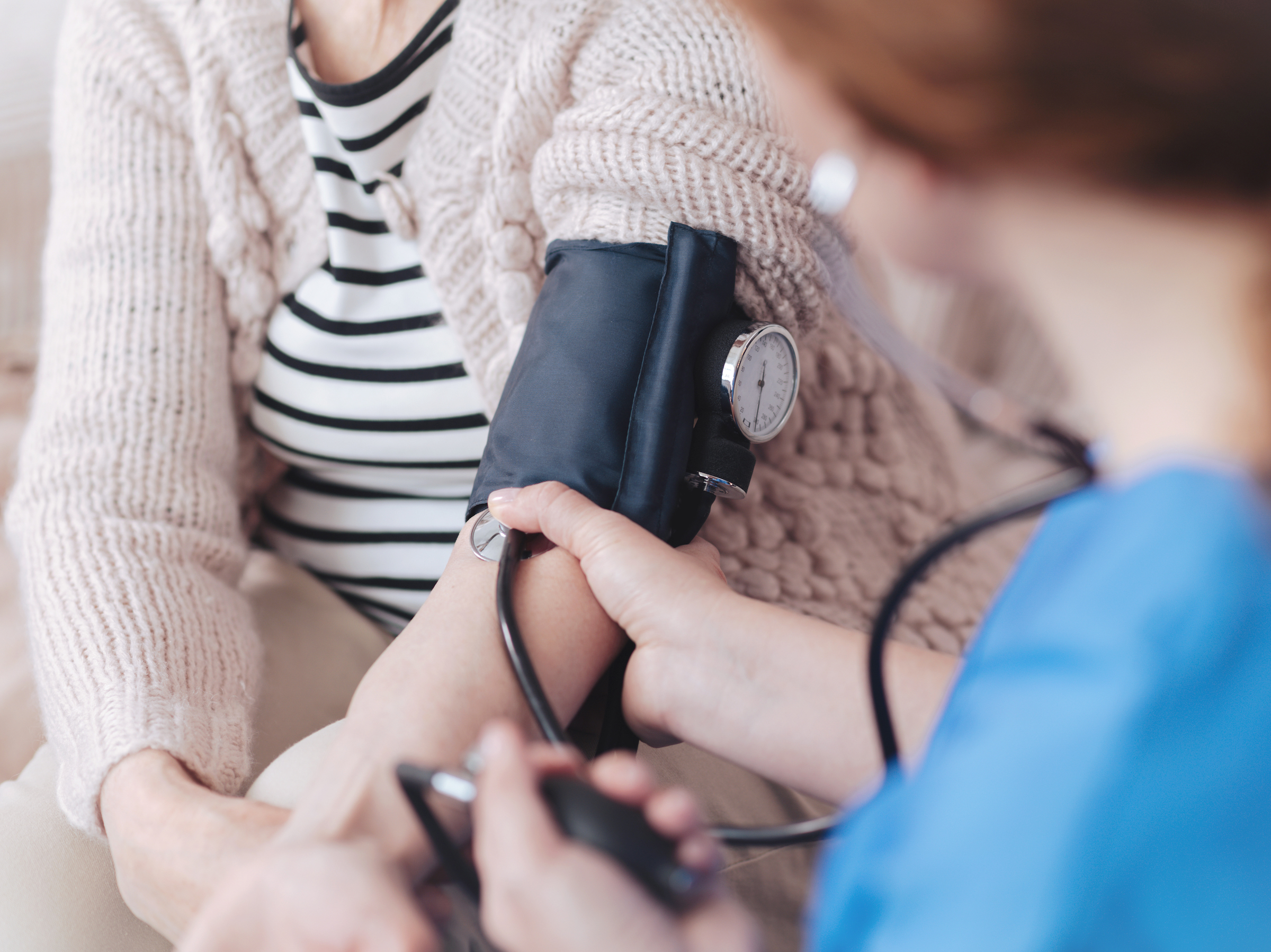 A nurse in blue takes the blood pressure of a woman wearing a striped shirt and sweater