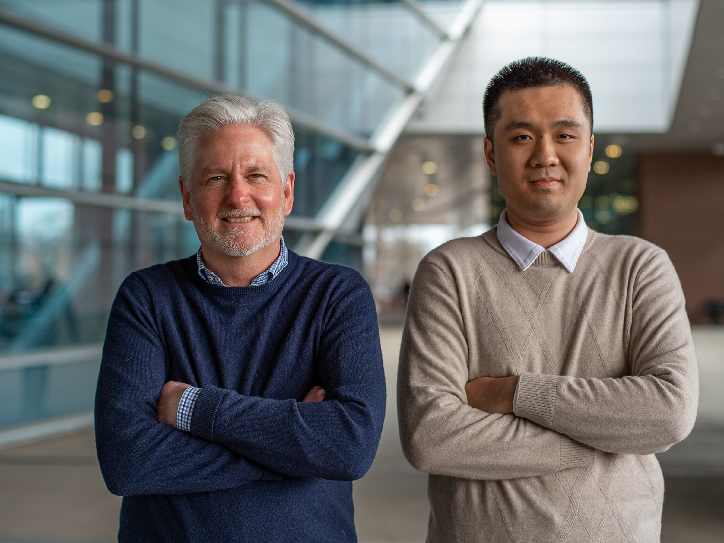 Two people stand with their arms crossed, facing the camera. Behind them, blue glass windows and a concrete path re visible.