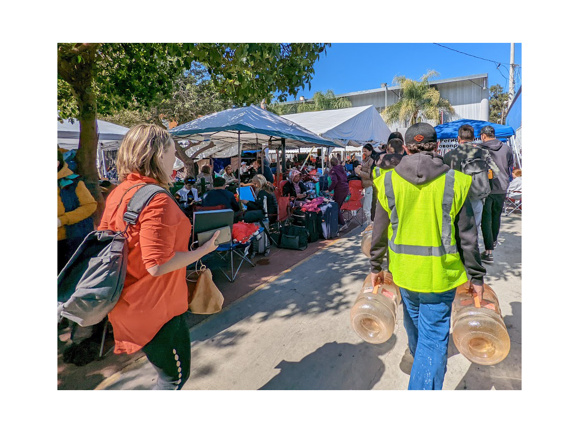 People gather underneath temporary canopies as a man wearing a neon yellow vest carries water jugs.