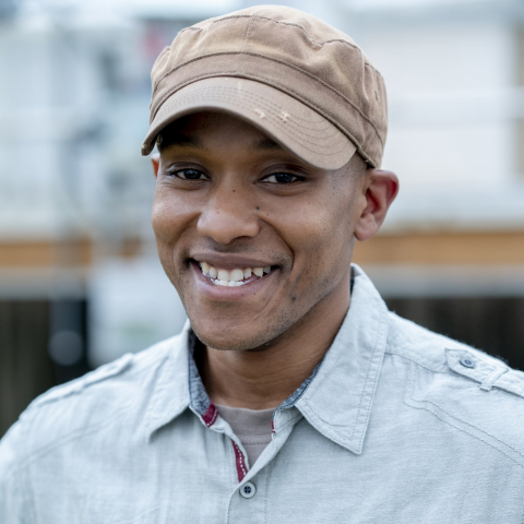 A man smiles at the camera wearing a brown hat and a blue collared shirt. 