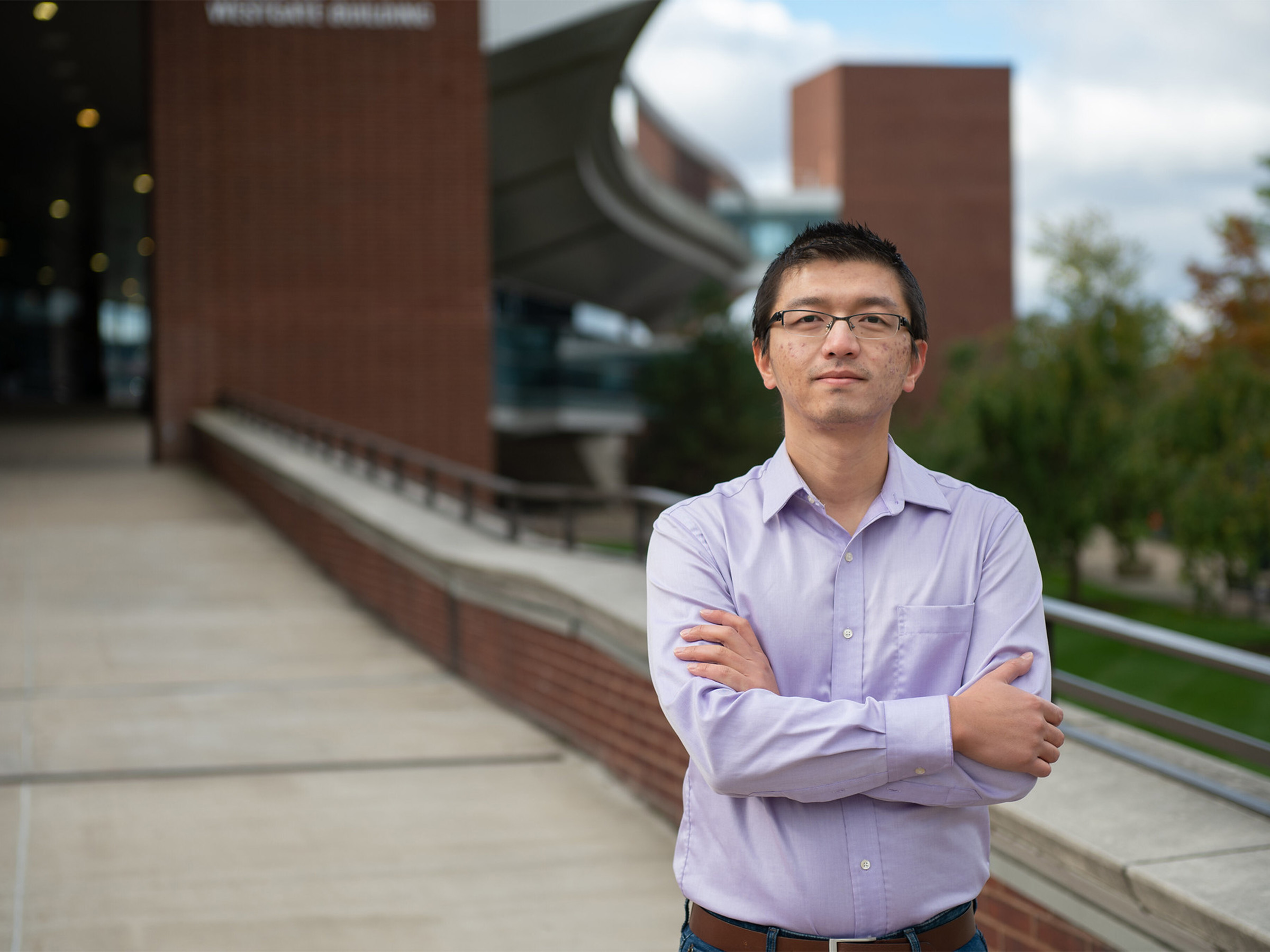 A man in a lavender shirt and glasses stands with his arms crossed on an outdoor walkway. 