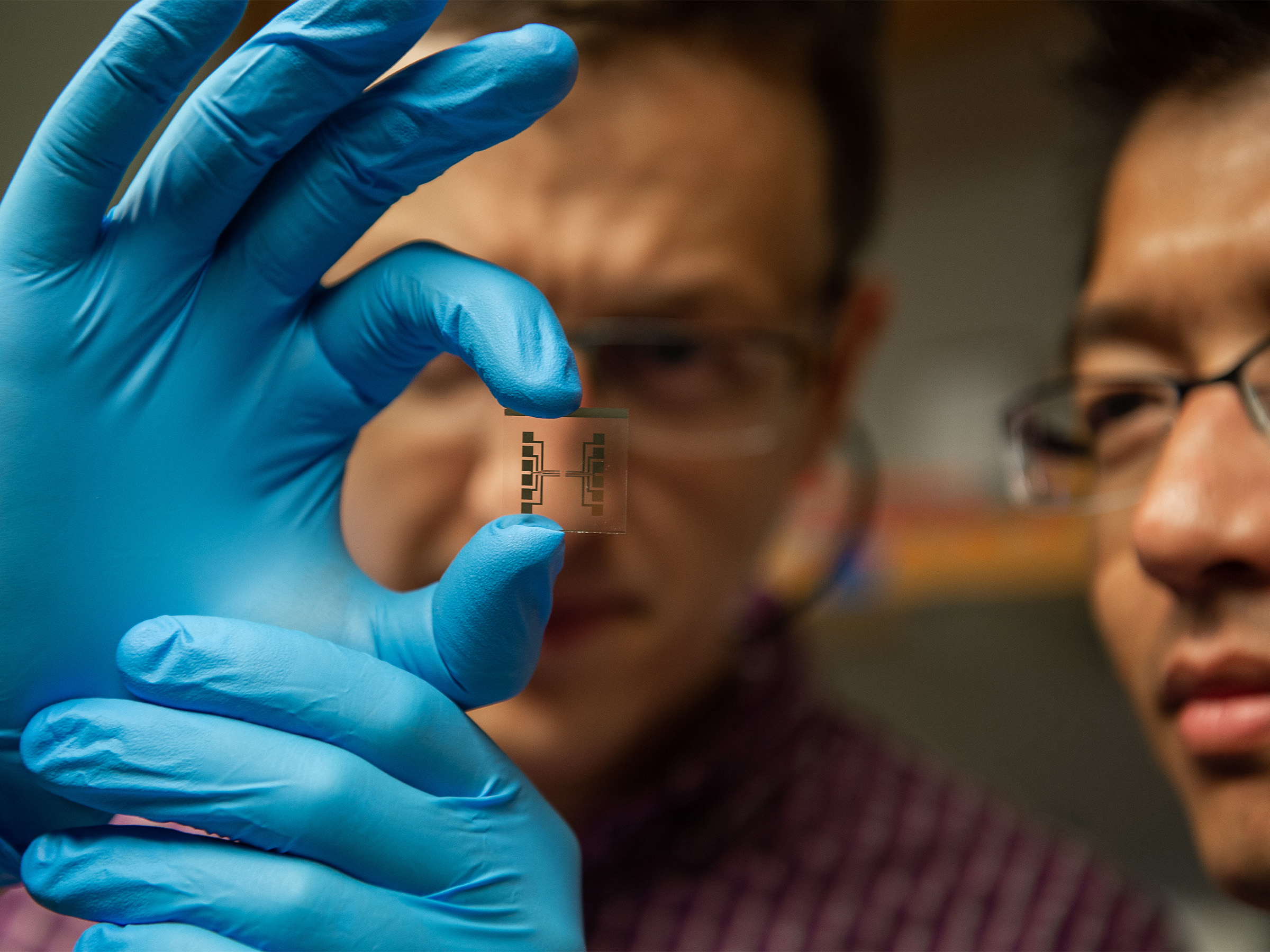 Hands wearing blue gloves hold up a transparent slide with black etching. Two faces are out of focus in the background.